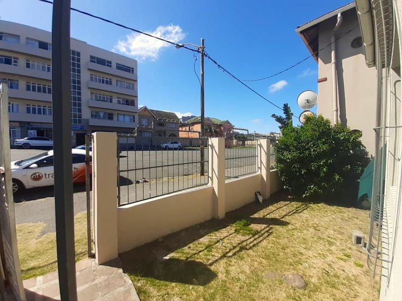 A rear exterior view of the property featuring a small lawn area, perimeter wall with metal security fencing, and building infrastructure.