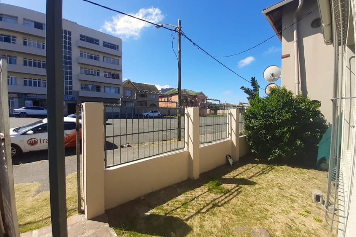 A rear exterior view of the property featuring a small lawn area, perimeter wall with metal security fencing, and building infrastructure.