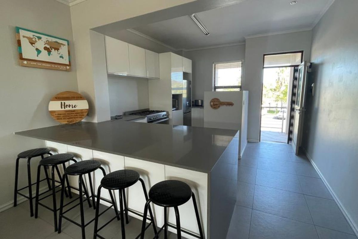 Modern open-plan kitchen featuring sleek white cabinetry, quartz countertops, a breakfast bar, and integrated gas cooking appliances, leading to an outdoor entrance.