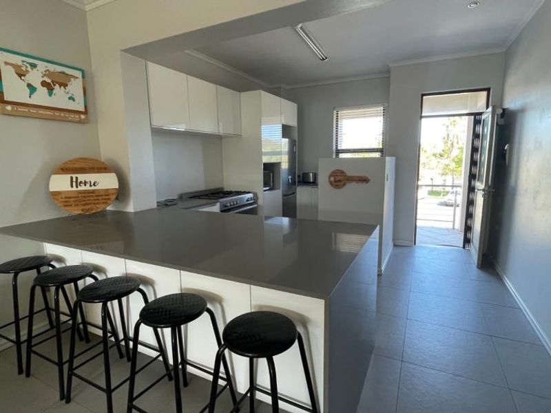 Modern open-plan kitchen featuring sleek white cabinetry, quartz countertops, a breakfast bar, and integrated gas cooking appliances, leading to an outdoor entrance.
