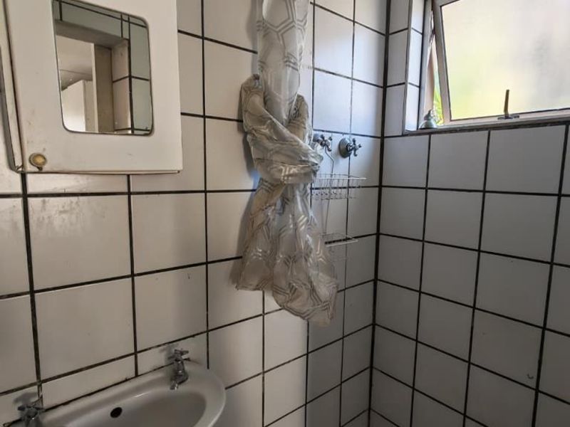 A compact bathroom featuring a wall-mounted basin, tiled shower enclosure, and a small medicine cabinet, requiring cosmetic updates to improve its dated appearance.