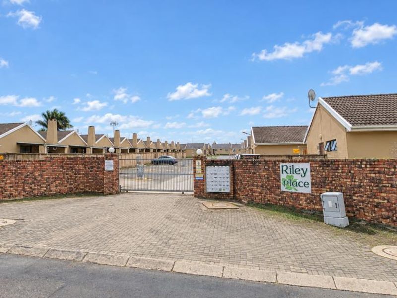Entrance view of Riley Place townhouse complex featuring a secure gated access point, brick boundary walls, and paved driveway.