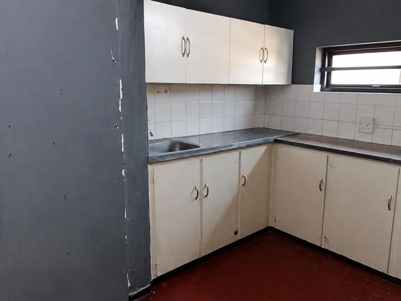 A compact, empty kitchen featuring built-in cabinetry, a stainless steel sink, and tiled backsplash, currently requiring cosmetic updates and wall repairs.