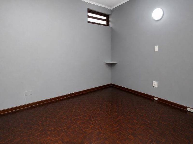 An empty bedroom featuring dark parquet flooring, grey walls, a corner shelf, and a wall-mounted light fixture.