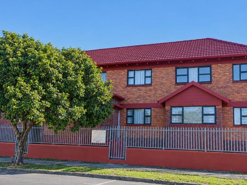 Front exterior view of a two-story brick apartment building featuring a red tiled roof, secure metal fencing, and a well-maintained brick boundary wall.