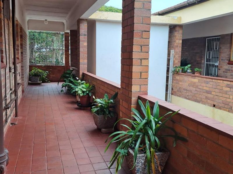 An open-air apartment balcony featuring brick pillars, a low brick balustrade, and tiled flooring, providing a functional outdoor walkway space.