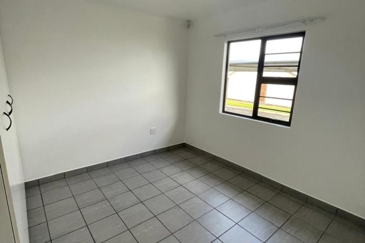 A clean, empty bedroom featuring neutral tiled flooring, a built-in cupboard, and a window fitted with security burglar bars.