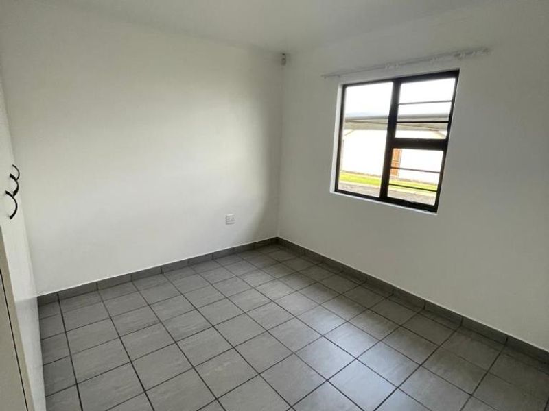 A clean, empty bedroom featuring neutral tiled flooring, a built-in cupboard, and a window fitted with security burglar bars.