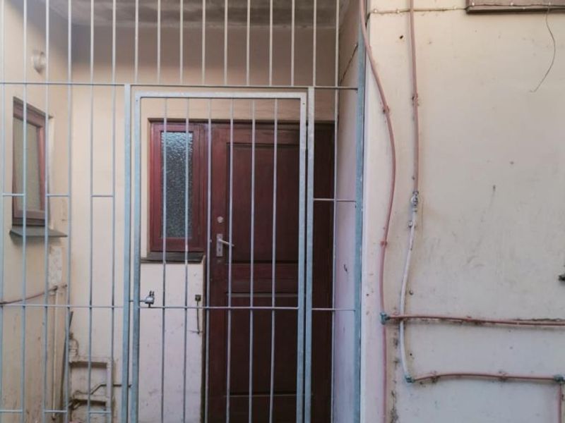 Exterior entrance area featuring a security gate, timber door, and window security bars, showing signs of wear on the surrounding wall surfaces.