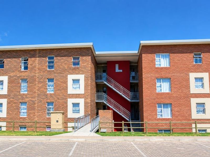 Front elevation of a multi-story brick apartment building featuring external staircases, paved parking, and secure perimeter fencing.