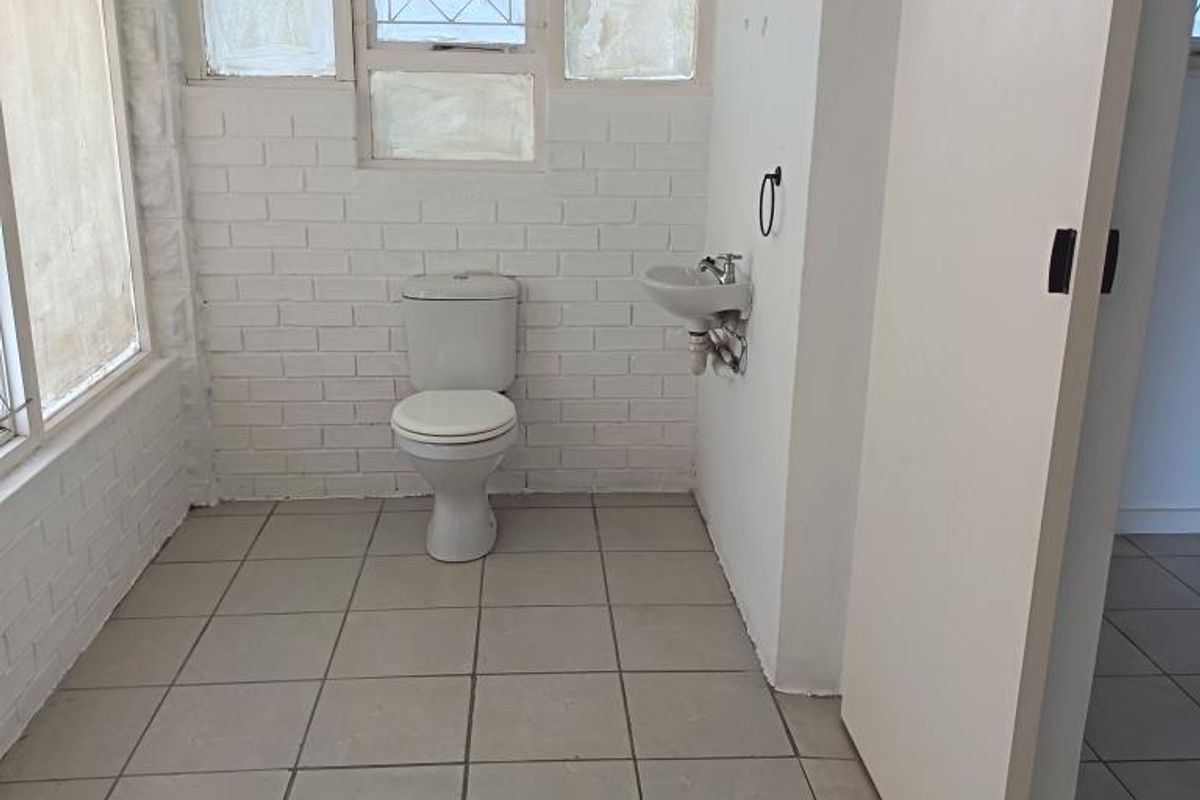 A functional bathroom featuring ceramic tile flooring, a wall-mounted basin, and a flush toilet, with exposed brick walls and natural light.