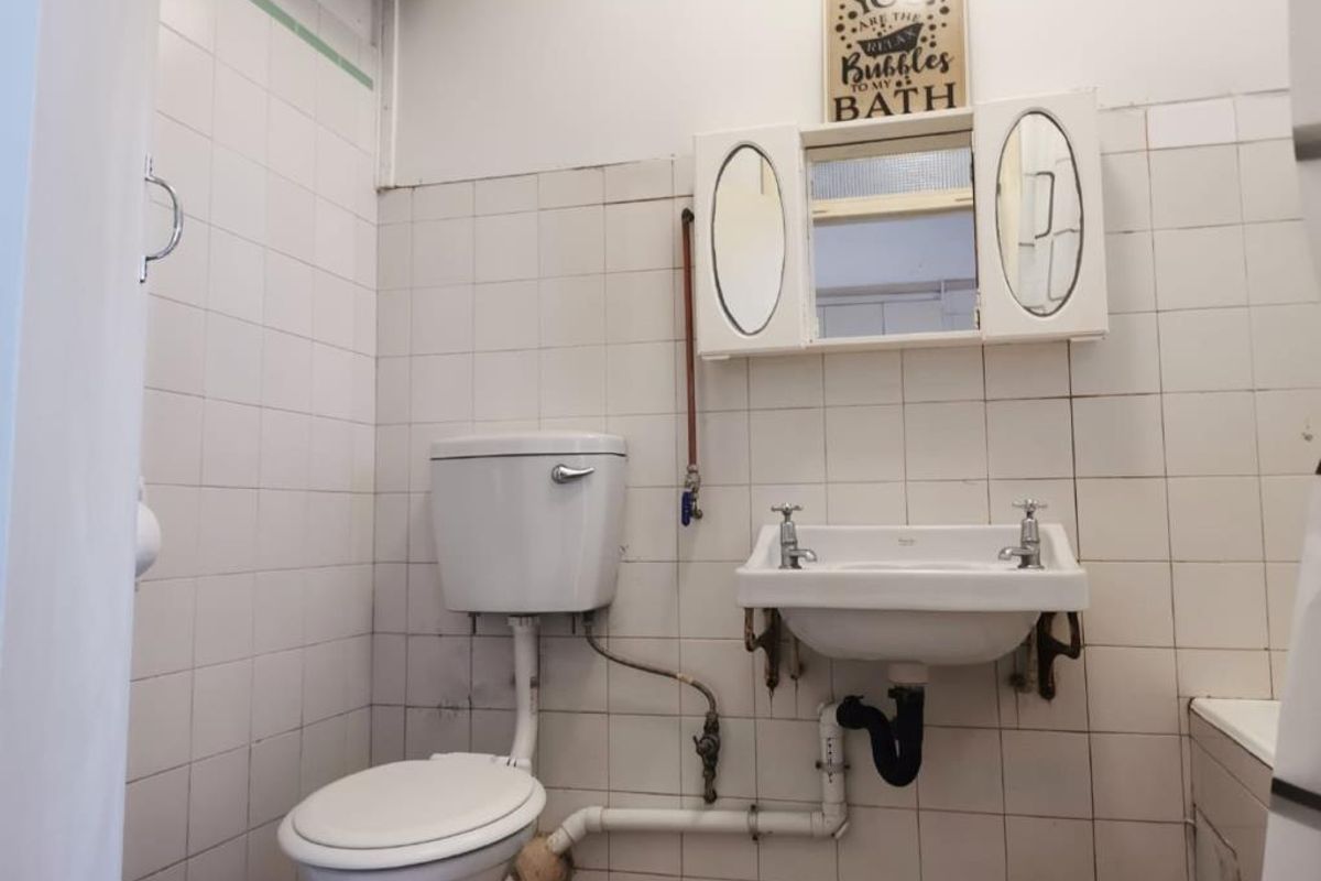 A dated bathroom featuring white wall tiles, a standard toilet, wall-mounted basin, and a medicine cabinet, requiring modernization of fixtures and finishes.