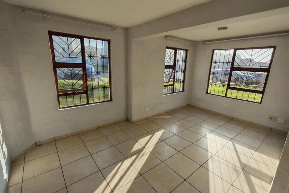 An empty, sunlit living area featuring tiled flooring, painted brick walls, and large windows fitted with security bars.