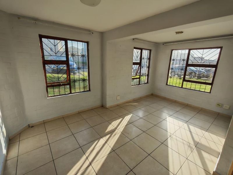 An empty, sunlit living area featuring tiled flooring, painted brick walls, and large windows fitted with security bars.