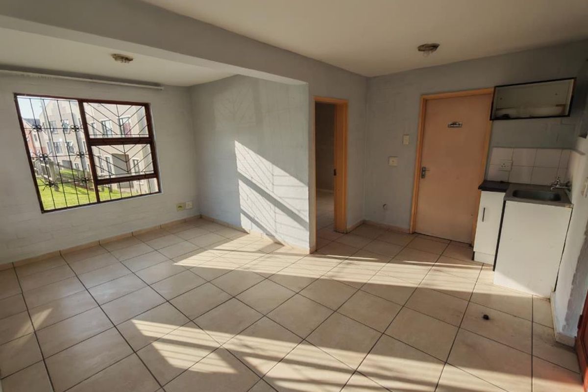 An open-plan living area featuring tiled flooring, a compact kitchen unit with a sink, and security bars on the window.
