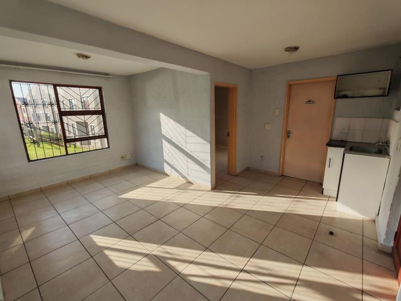 An open-plan living area featuring tiled flooring, a compact kitchen unit with a sink, and security bars on the window.