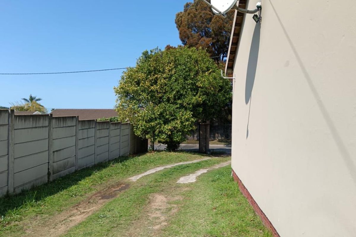 A side exterior view of the property featuring a grass driveway, concrete boundary wall, and exterior wall-mounted satellite dish.
