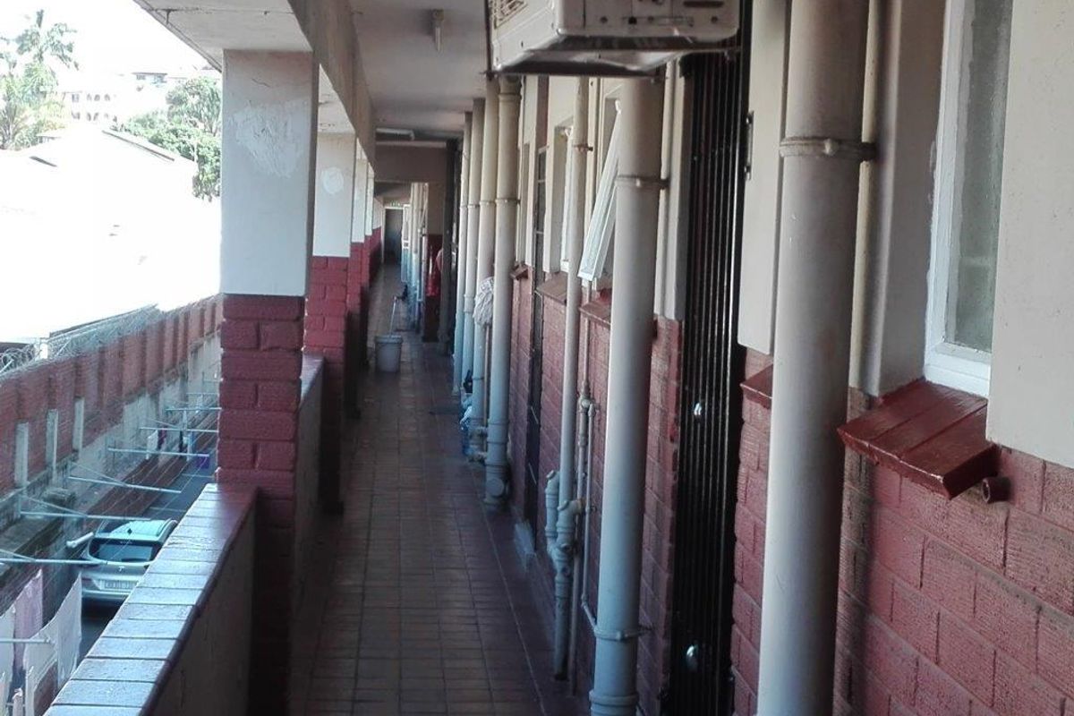 An exterior apartment building walkway featuring tiled flooring, exposed plumbing, and security gates, showing signs of wear and aging.