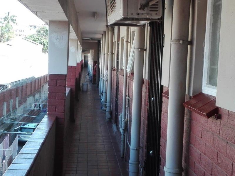 An exterior apartment building walkway featuring tiled flooring, exposed plumbing, and security gates, showing signs of wear and aging.