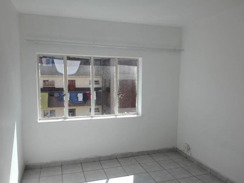 An empty bedroom featuring tiled flooring and a large metal-framed window with a curtain rail, offering a view of the adjacent building.