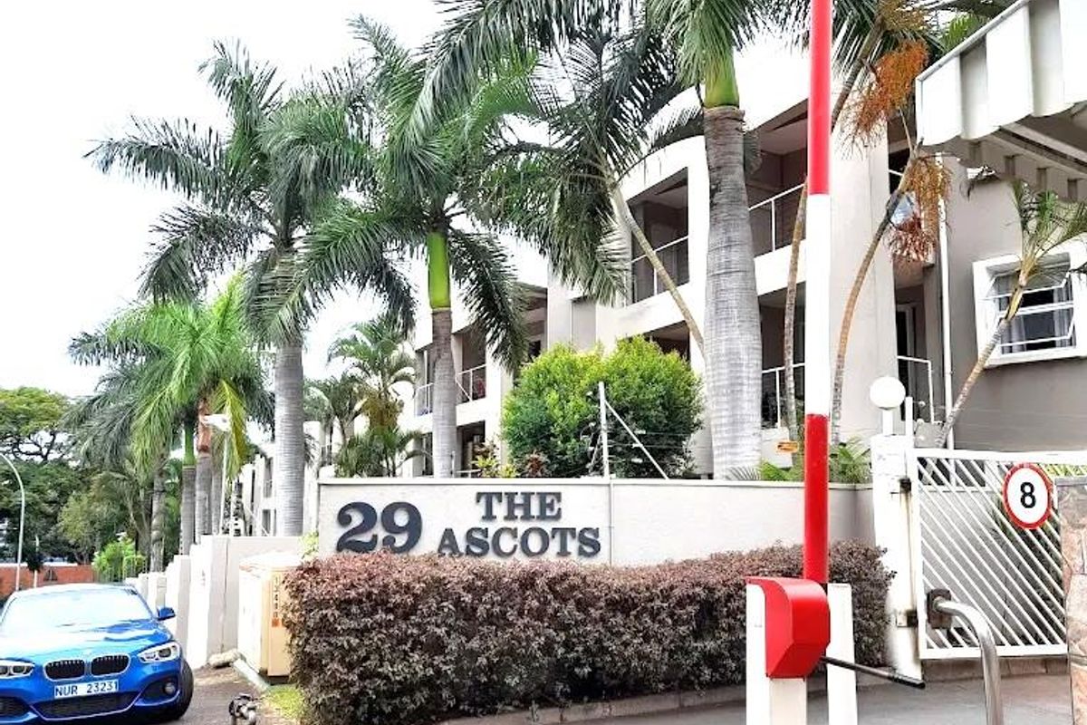 The entrance to The Ascots apartment complex, featuring a security boom gate, signage, and established palm trees along the driveway.