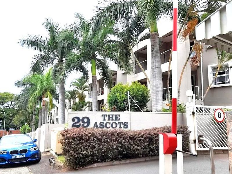 The entrance to The Ascots apartment complex, featuring a security boom gate, signage, and established palm trees along the driveway.