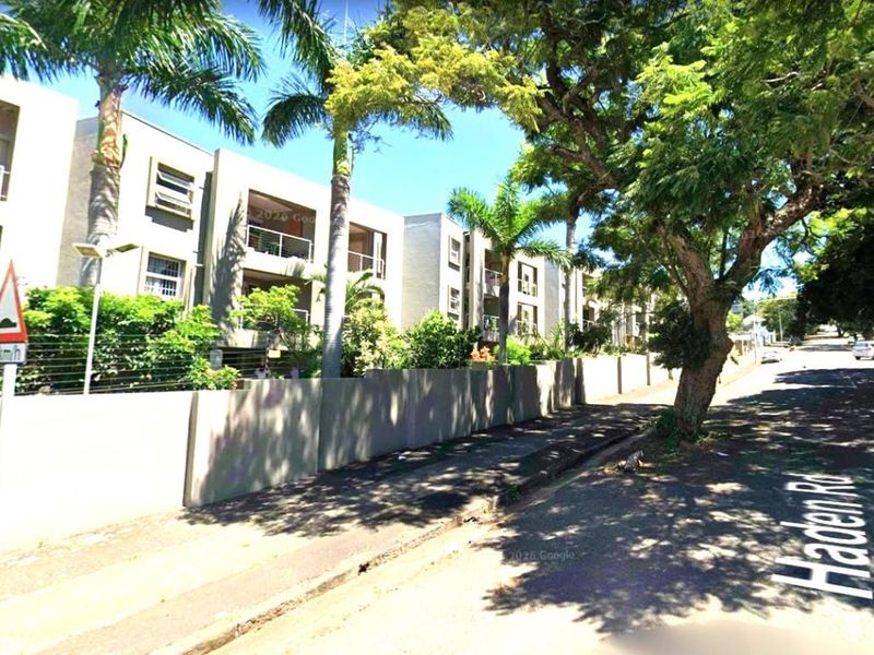 Street view of the apartment complex exterior, featuring multi-story residential units with balconies, perimeter walling, and mature landscaping along the roadside.