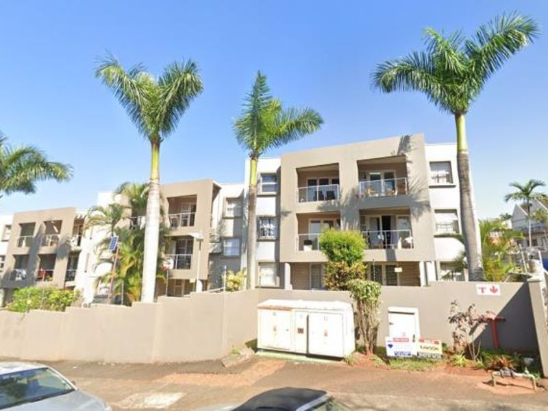 Street-level view of a modern multi-story apartment complex featuring balconies, perimeter walling, and established palm tree landscaping.