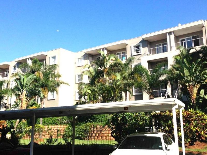 Exterior view of a multi-story apartment complex featuring private balconies, a covered carport, and landscaped garden areas.