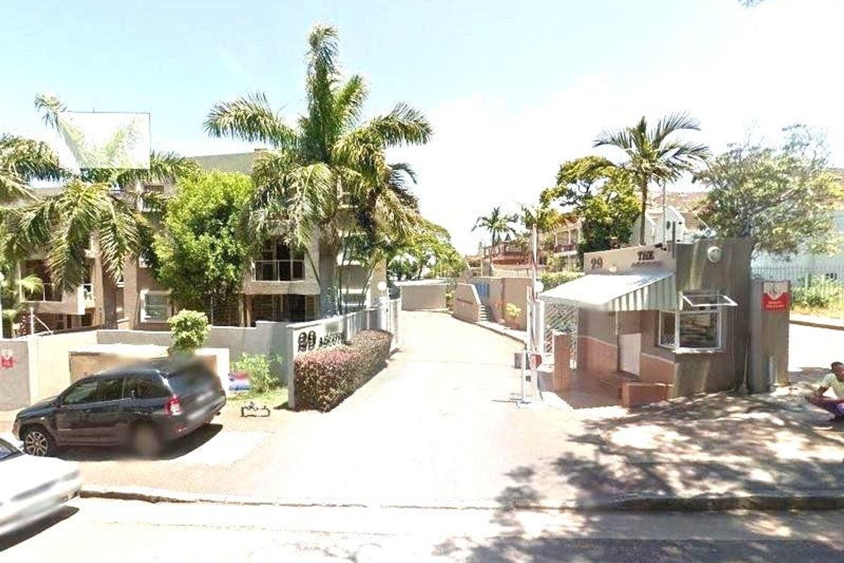 Street view of the apartment complex entrance featuring a security gatehouse, perimeter wall, and paved driveway access.