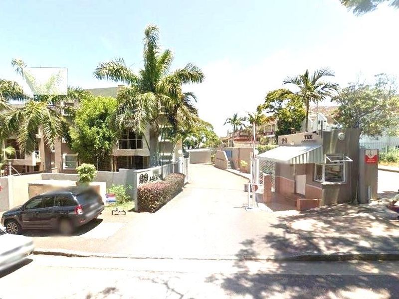 Street view of the apartment complex entrance featuring a security gatehouse, perimeter wall, and paved driveway access.