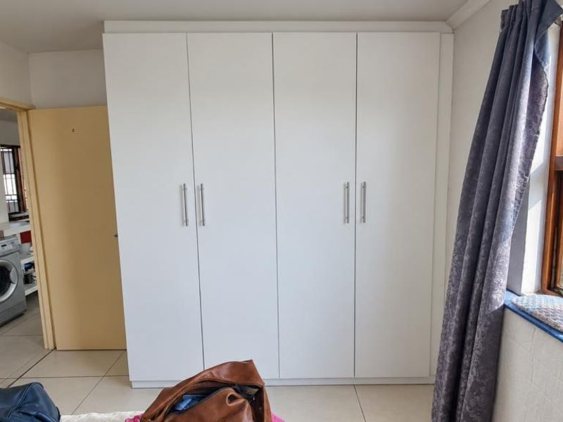 A bedroom featuring a large white built-in wardrobe, tiled flooring, and a timber-framed window with natural light.