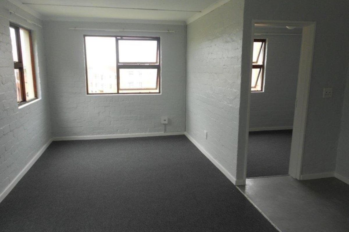 A vacant bedroom featuring grey carpeting, painted brick walls, and multiple windows providing natural light, with an adjacent room visible through the doorway.