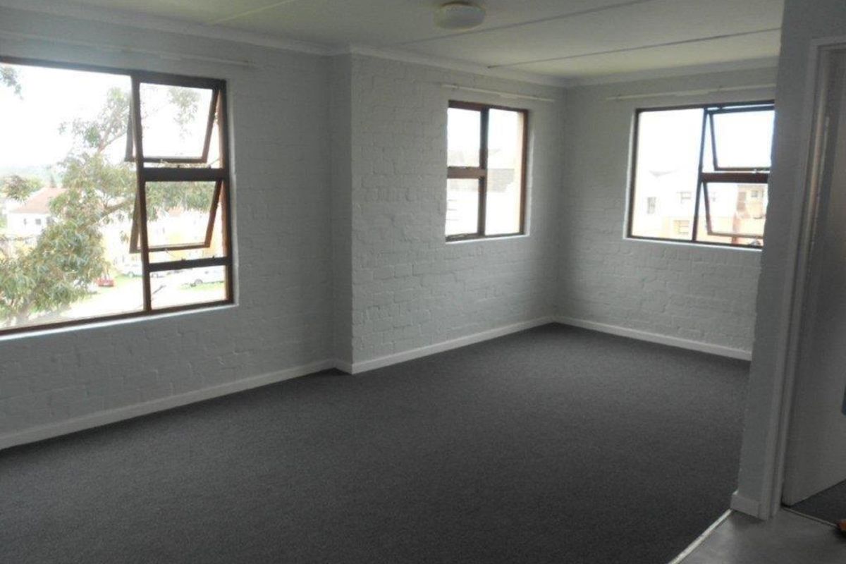 A spacious, empty living area featuring painted brick walls, grey carpeting, and large windows providing natural light.