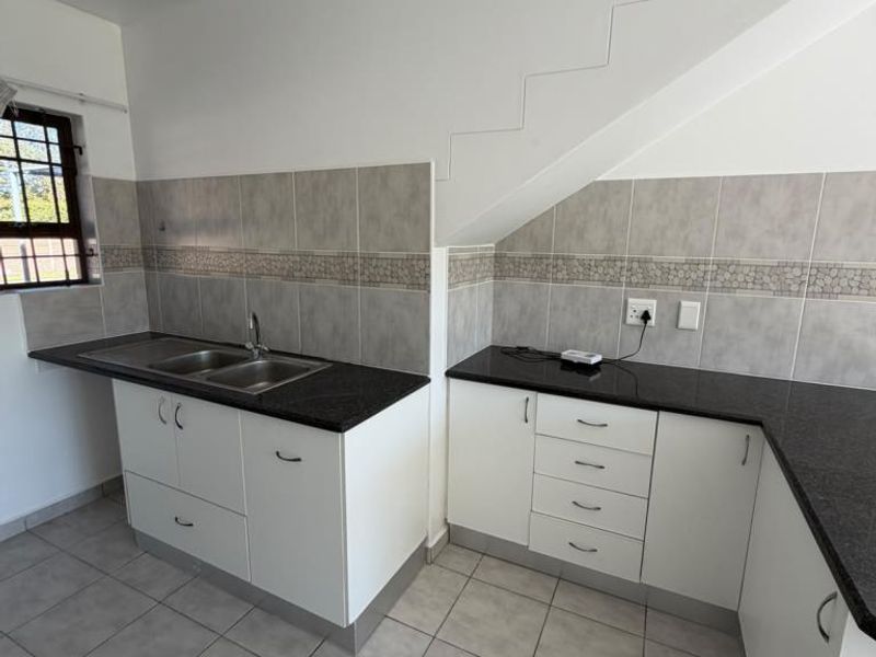 A clean, empty kitchen featuring white cabinetry, dark granite countertops, double sink, and neutral ceramic tiling, situated under a staircase.