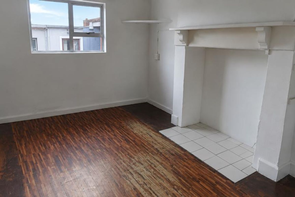 An empty living room featuring a traditional fireplace mantel, tiled hearth, and original parquet flooring in need of restoration.