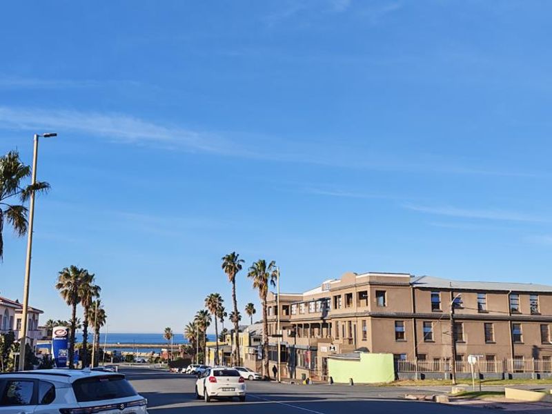 A street-level view of the neighbourhood approach, showing surrounding buildings, palm trees, and a glimpse of the ocean in the distance.