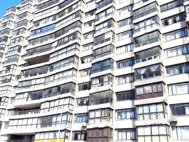 A low-angle view of a large multi-story apartment building featuring numerous enclosed balconies and a ground-level entrance canopy.