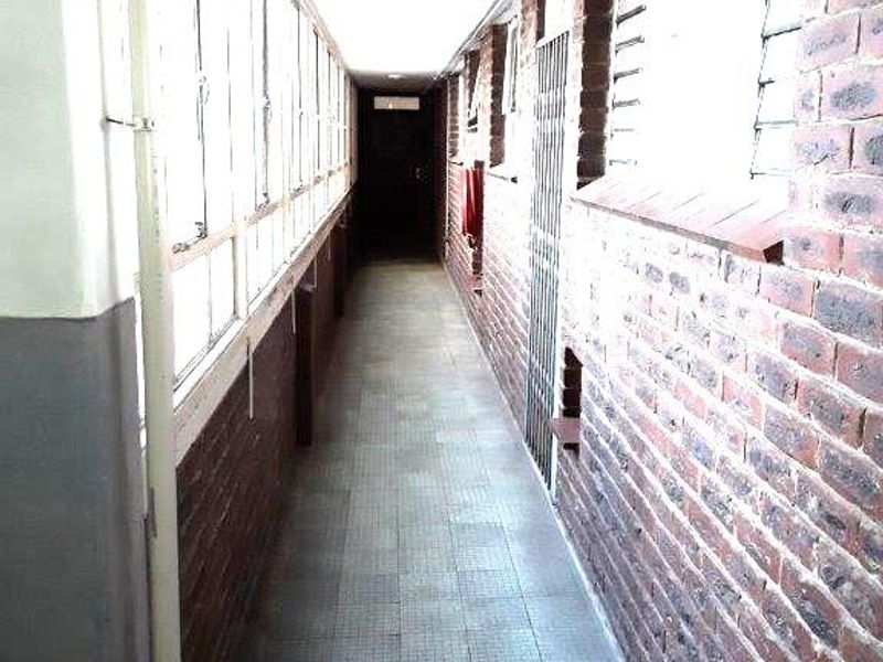 A long, narrow hallway featuring exposed brick walls, tiled flooring, and large windows, providing access to the apartment's interior spaces.
