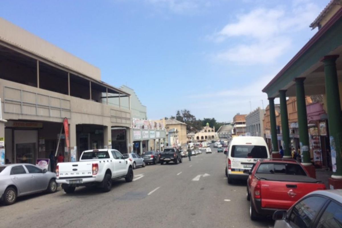 A street-level view of a commercial district featuring storefronts with covered walkways and structural columns along a busy road.