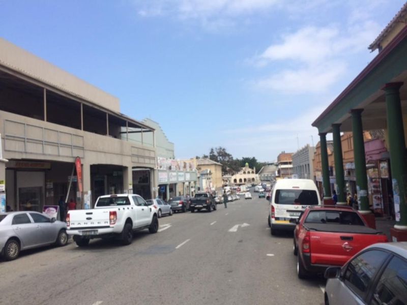 A street-level view of a commercial district featuring storefronts with covered walkways and structural columns along a busy road.