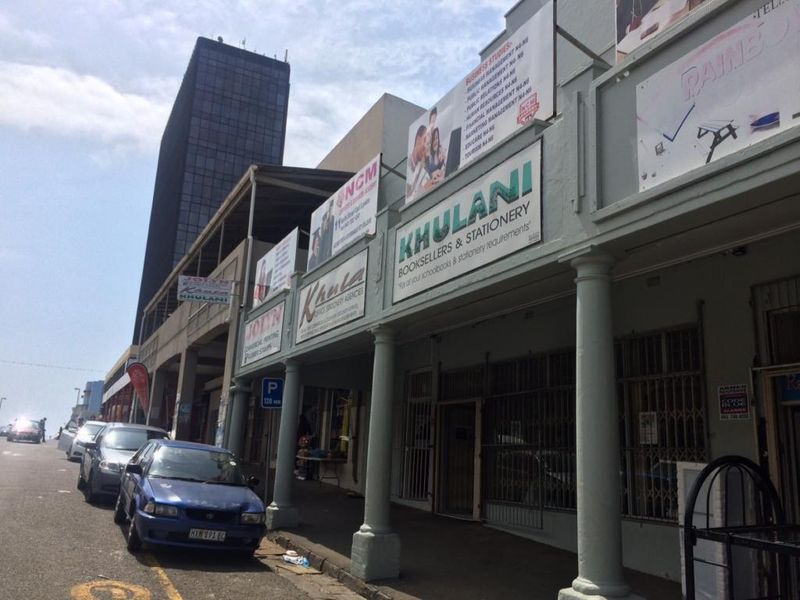 Street-level view of a commercial building facade featuring multiple business signs, security gates, and structural columns along the sidewalk.