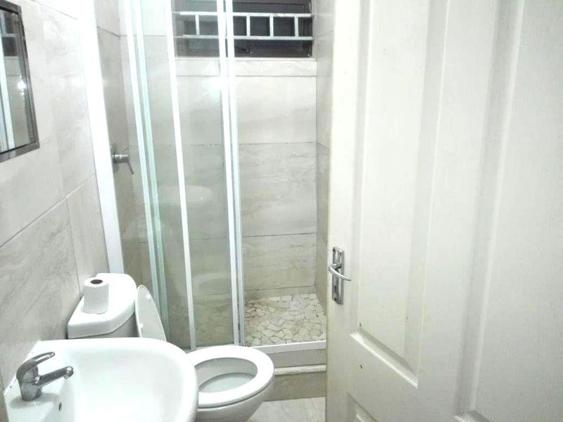 Compact bathroom featuring a tiled shower enclosure, pedestal basin, and toilet, finished with neutral wall tiles and a small high-level window.