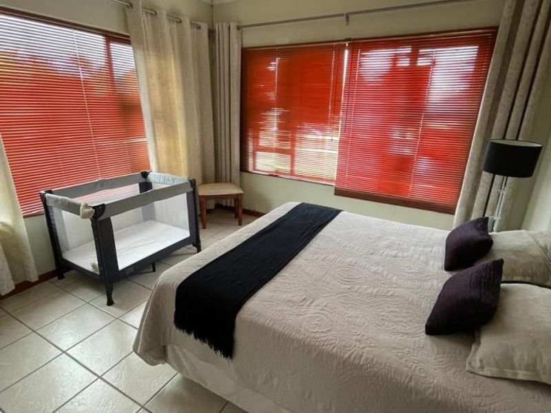 A bedroom featuring tiled flooring, a ceiling fan, and large windows with venetian blinds, offering a functional space with natural light.