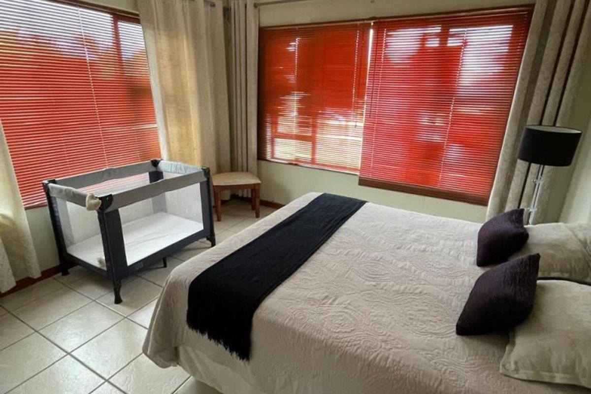 A bedroom featuring tiled flooring, a ceiling fan, and large windows with venetian blinds, offering a functional space with natural light.