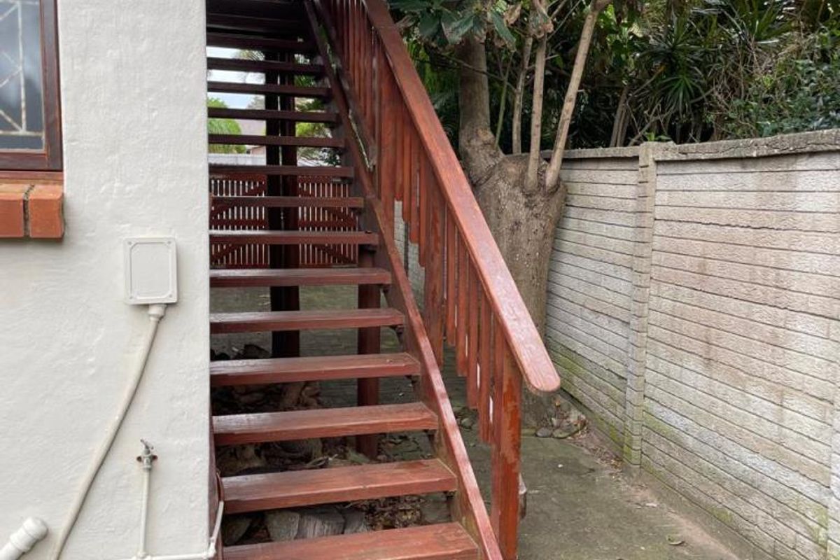 Rear exterior view showing a wooden staircase leading to an upper level, set against a concrete boundary wall and paved ground area.