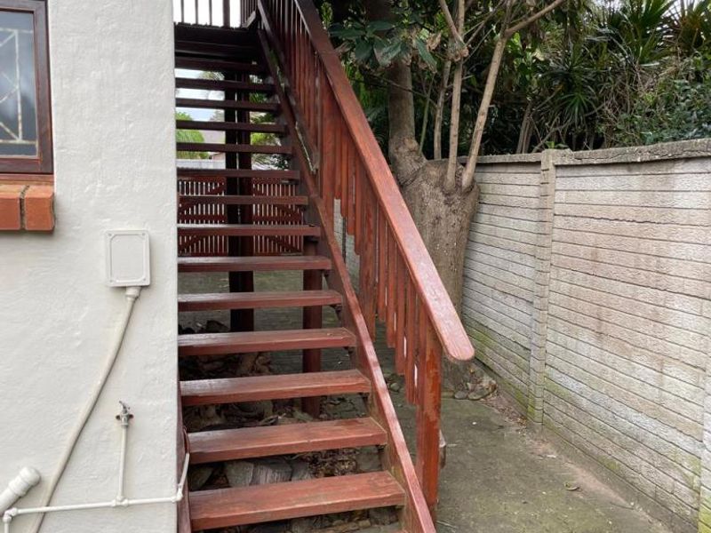 Rear exterior view showing a wooden staircase leading to an upper level, set against a concrete boundary wall and paved ground area.
