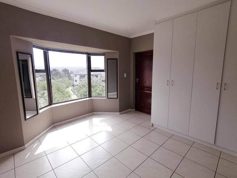 A bright, empty bedroom featuring tiled flooring, built-in cupboards, and a large bay window offering natural light and scenic views.