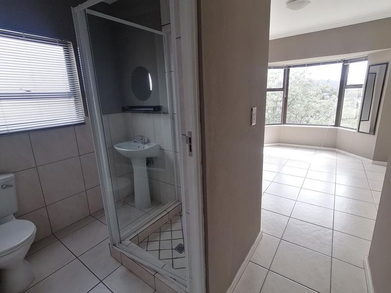 A bright, empty bathroom featuring a shower enclosure, pedestal basin, and toilet, adjacent to a spacious room with large bay windows.