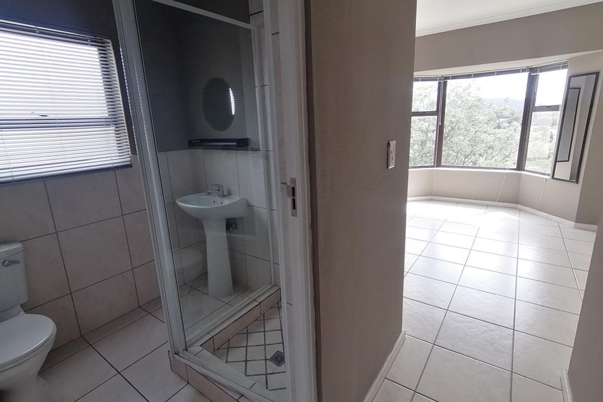 A bright, empty bathroom featuring a shower enclosure, pedestal basin, and toilet, adjacent to a spacious room with large bay windows.